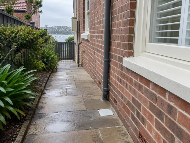 Outdoor brick side path with a covered drain beside wet paving and a house wall in Clovelly